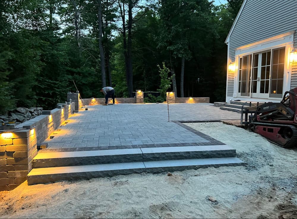 Patio construction in progress at night with lights, surrounded by trees and a house.